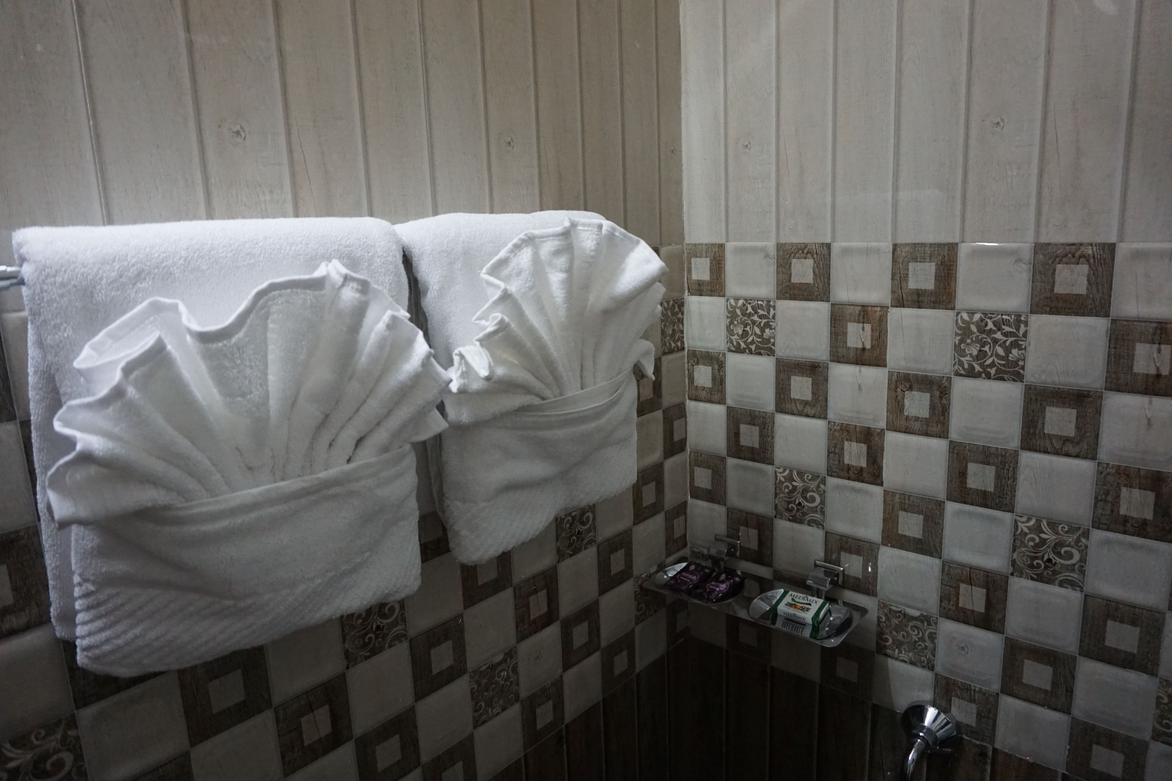 A bathroom with two white towels hanging on a rack, a small shelf with toiletries, and a tiled wall with a checkered pattern.