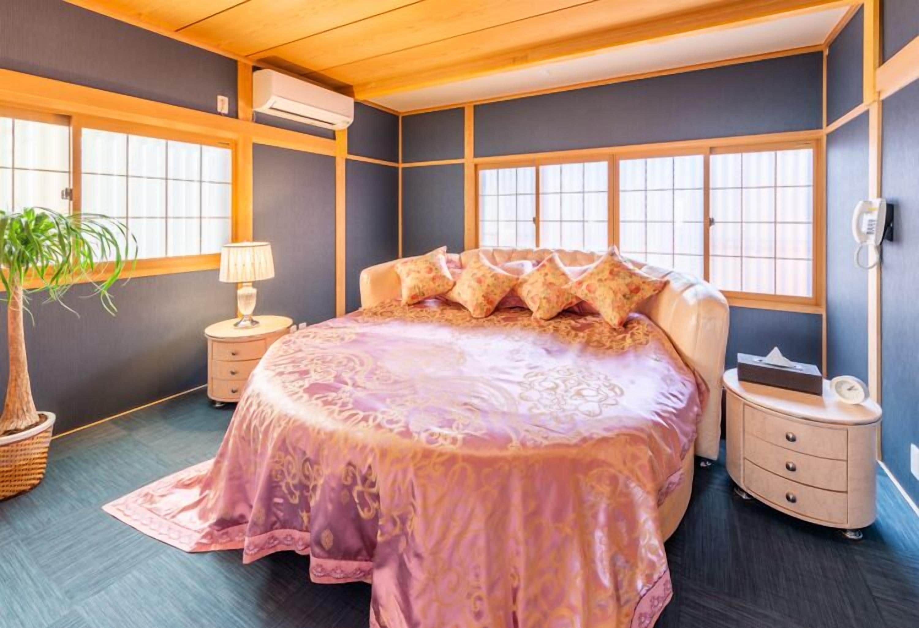 Une chambre à coucher avec un grand lit, une table de chevet, une lampe, une plante et une fenêtre avec des stores.