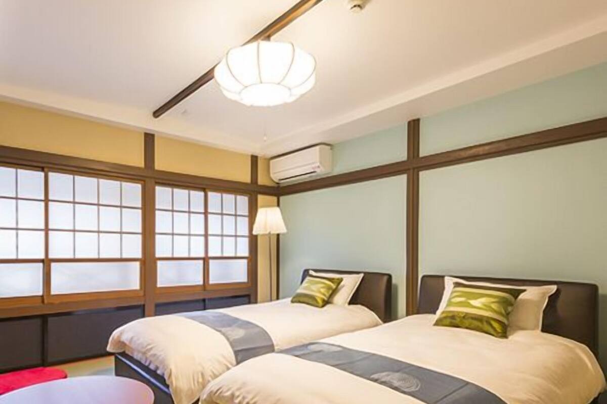 A hotel room with two beds, a small table, a chair, and a window with a view.