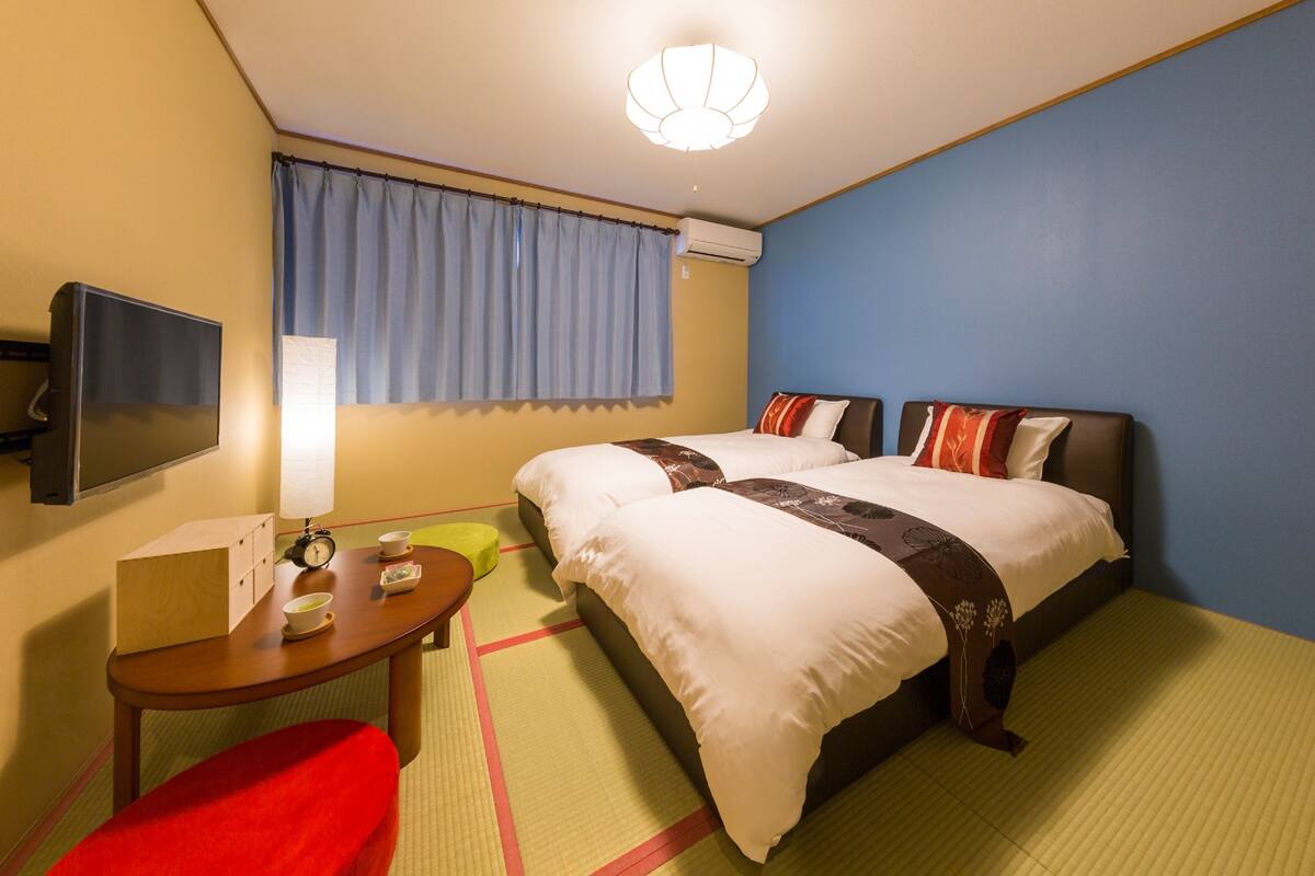 A hotel room with two beds, a TV, a table with a box and a clock, and a window with curtains.