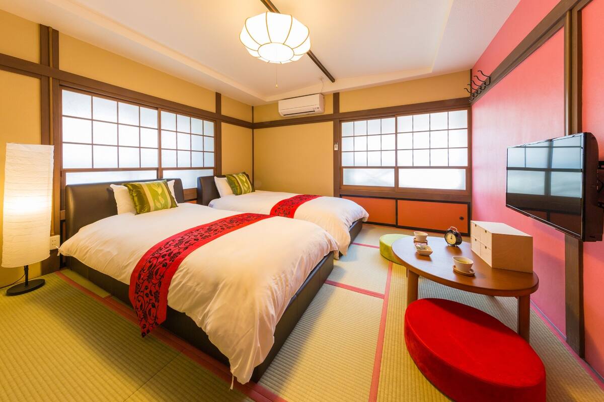 A hotel room with two beds, a TV, a table with a clock, and a window with a view.