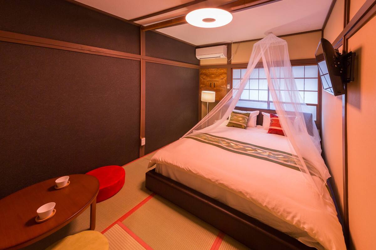 A hotel room with a bed, a small table with cups, a red stool, and a TV mounted on the wall.