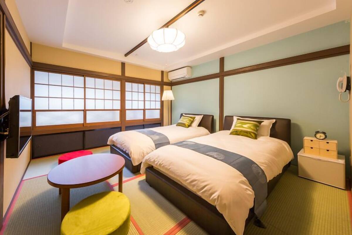 A hotel room with two beds, a small table, a red stool, and a nightstand with a clock.