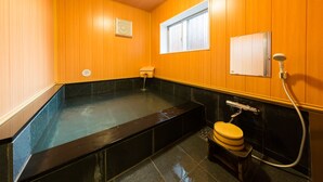 Un onsen japonés tradicional con interior de madera, una zona de remojo negra, una ventana pequeña y un inodoro con ducha.