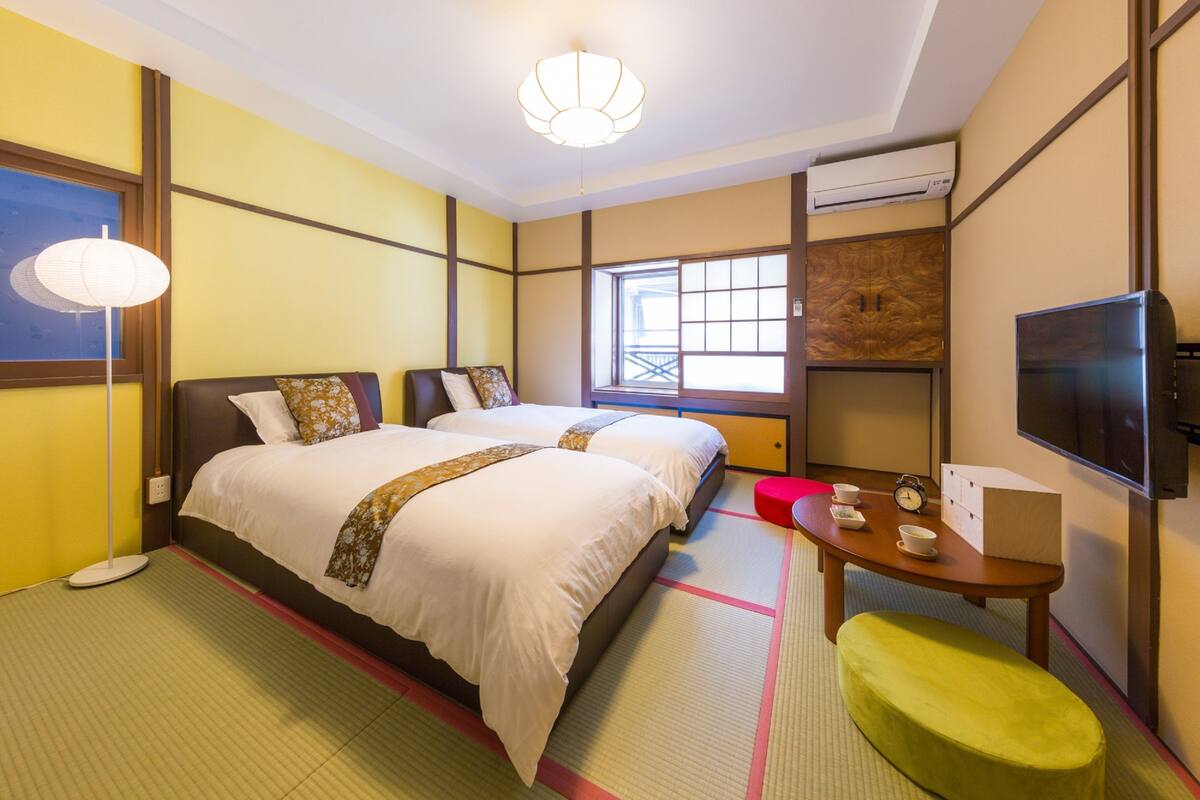A hotel room with two beds, a TV, a table with a clock and cups, and a window with a view.