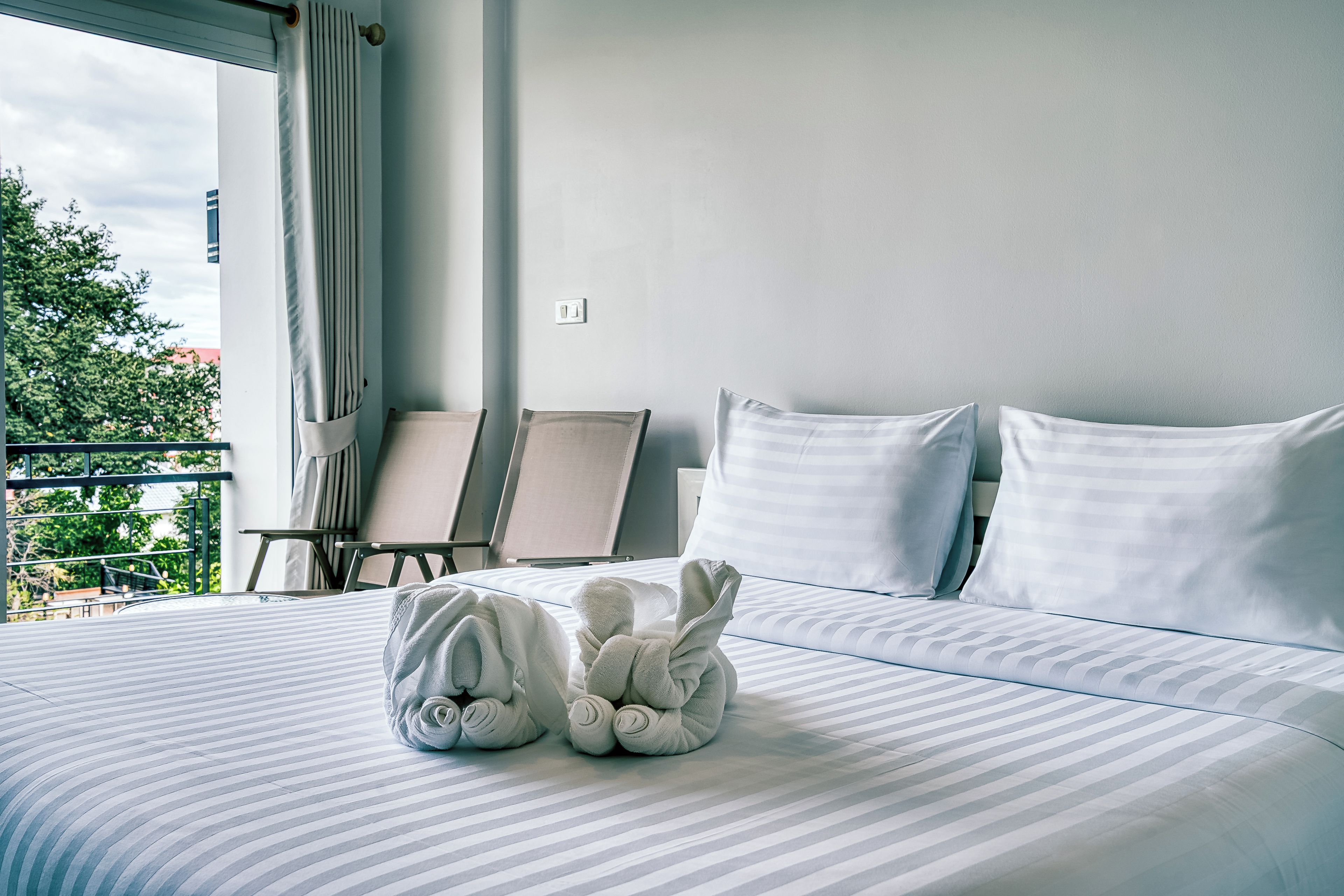 Habitación de hotel con cama, dos almohadas, un par de toallas, ventana con cortinas y vista a zonas verdes.
