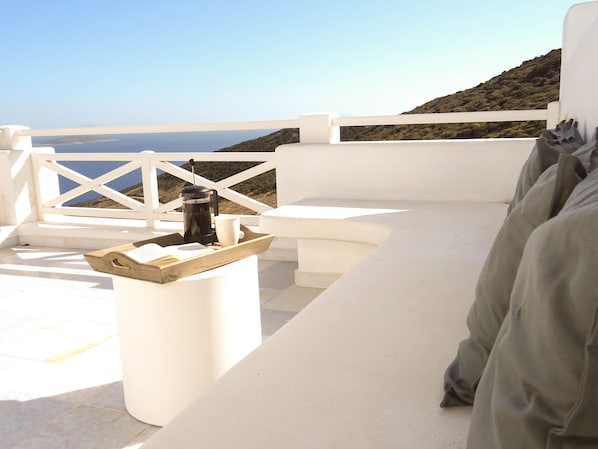 Zona de estar al aire libre con mobiliario blanco, una cafetera y una taza, con vistas a un cuerpo de agua y colinas.