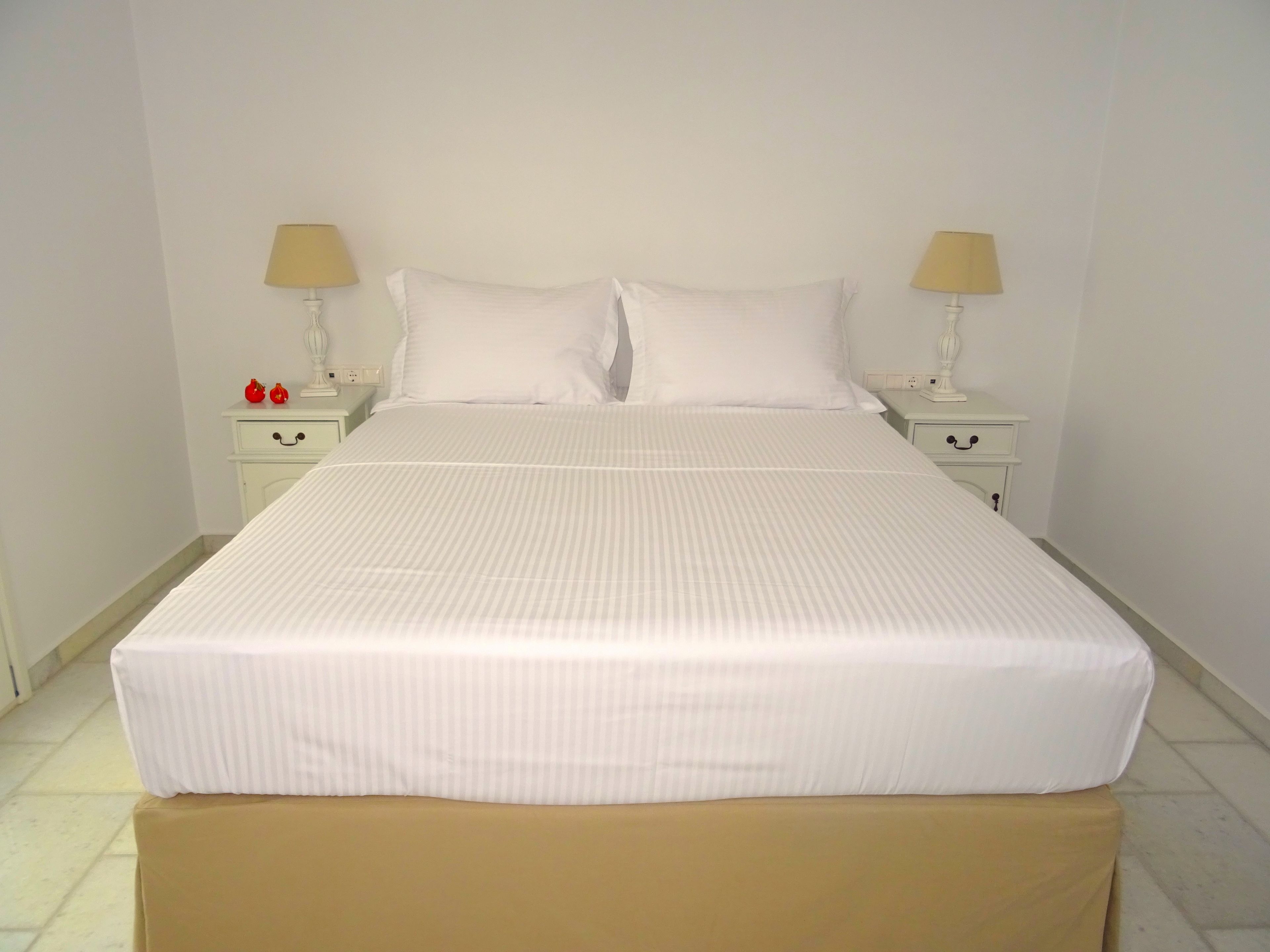 A neatly made bed with white bedding, two bedside tables with lamps, and a small drawer unit.
