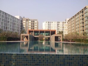 A modern hotel with a central building surrounded by a swimming pool, reflecting the nearby high-rise buildings. - D Condo Sign Chiang Mai R628 (Chiang Mai)