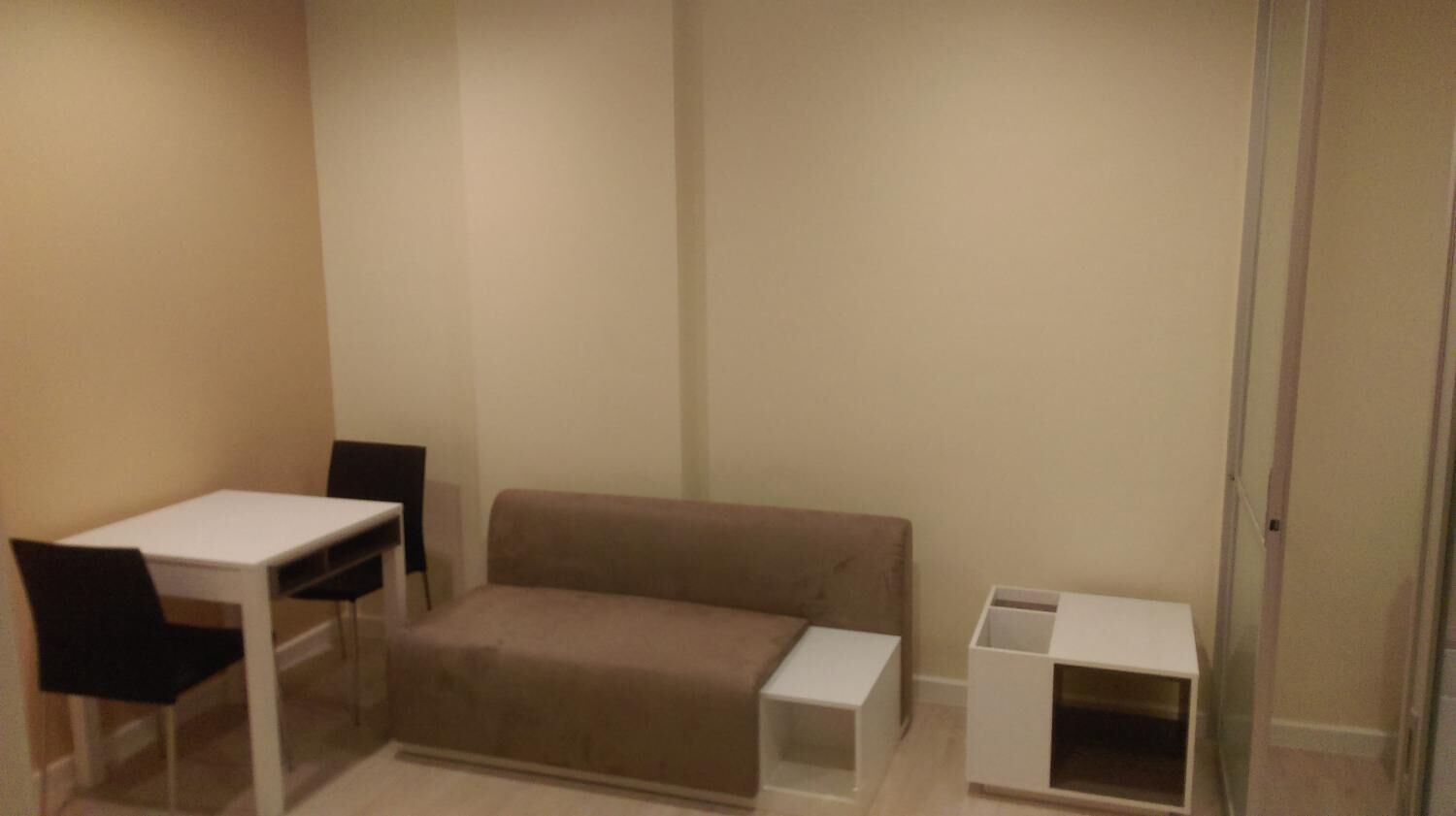 A room with a beige sofa, a white table, and two black chairs. There is a white cabinet with a shelf.