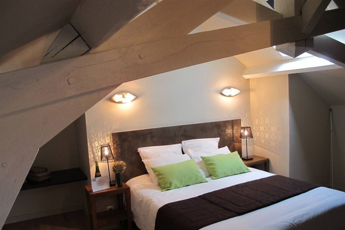 A bedroom with a sloped ceiling, a bed with white and green bedding, two bedside lamps, and a small table with a vase.