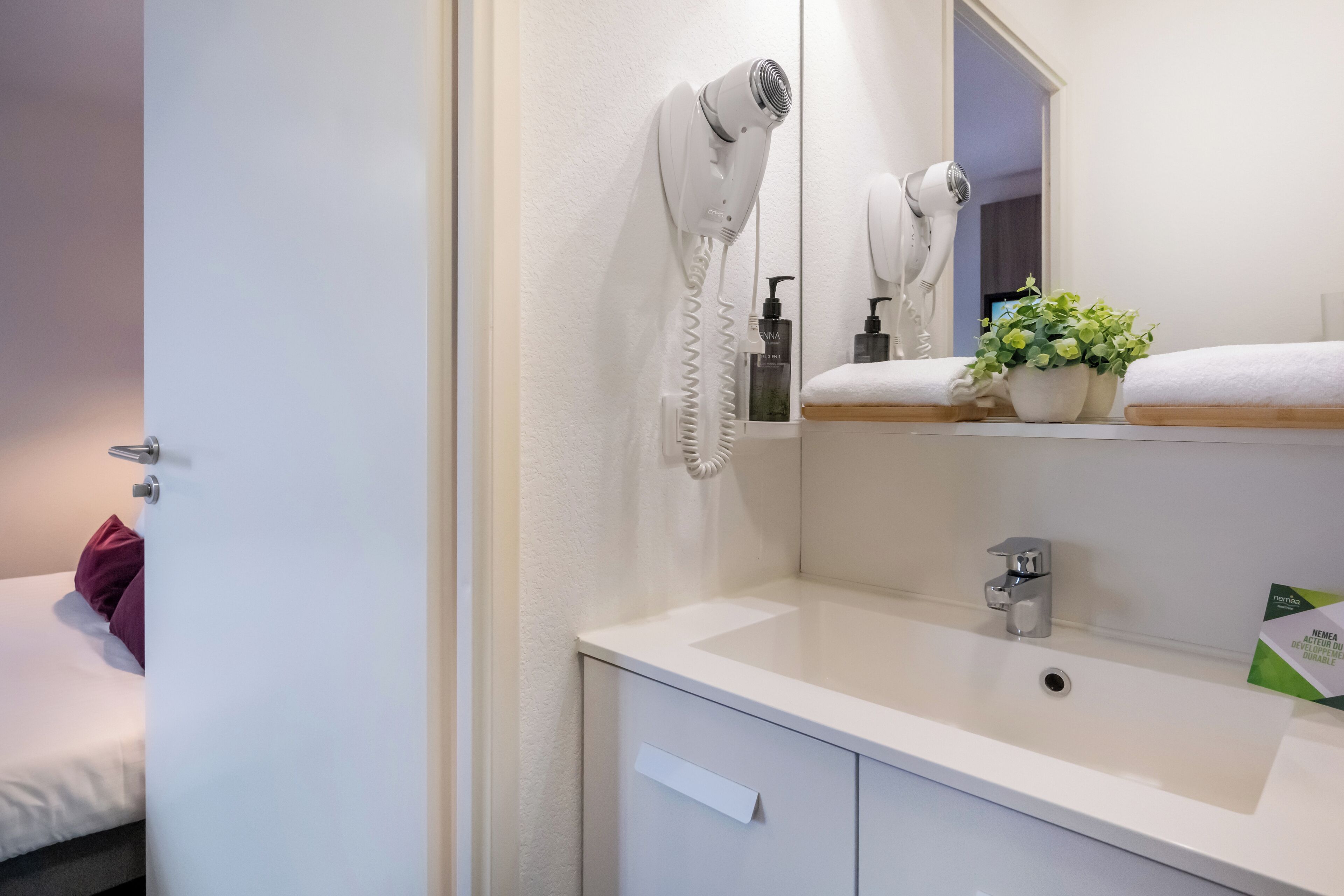 Casa de banho moderna com lavatório branco, espelho, secador de cabelo, uma planta em vaso e toalha sobre o balcão.