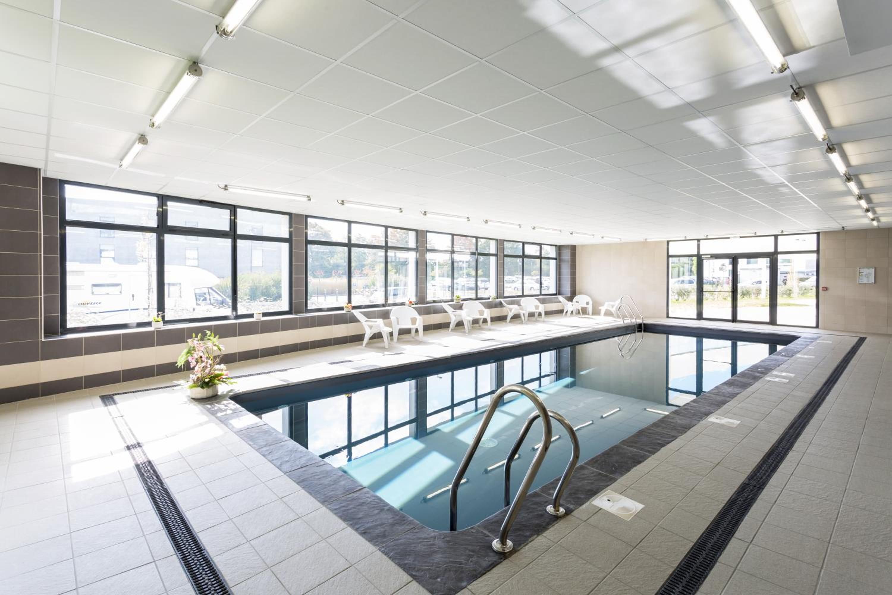 Une grande piscine intérieure avec de larges baies vitrées, un sol carrelé et quelques chaises disposées le long du bord.