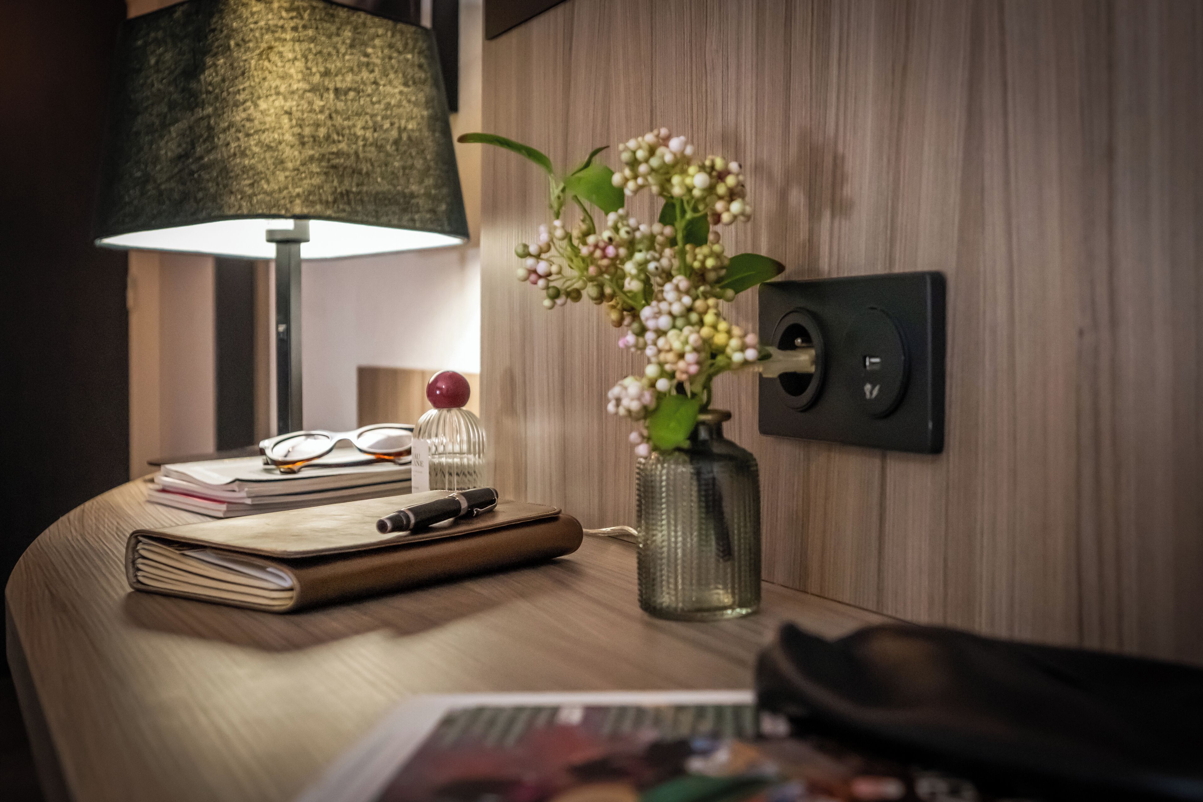 A neatly arranged desk with a lamp, a vase with flowers, a notebook, glasses, and a pen.