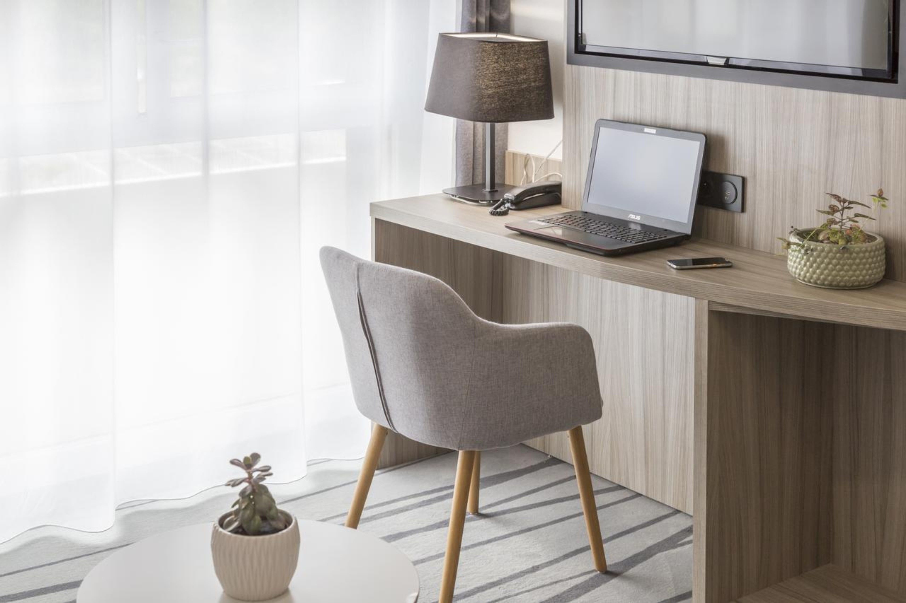 A modern hotel room with a wooden desk, a laptop, a lamp, a phone, and a chair.