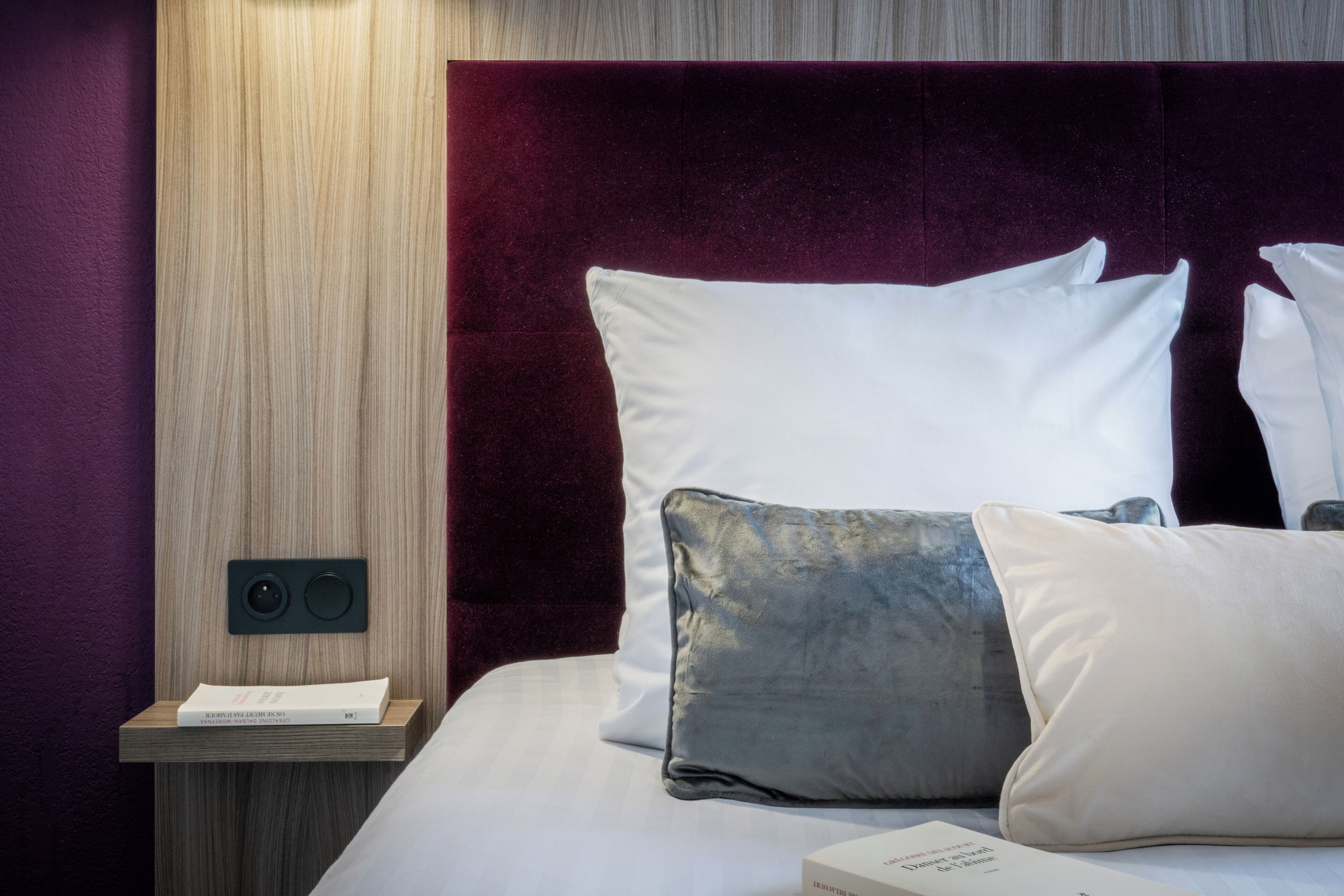 A neatly made bed with white and grey pillows, a wooden bedside table with a book, and a dark red headboard.