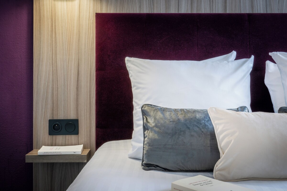 A neatly made bed with white and grey pillows, a wooden bedside table with a book, and a dark red headboard.