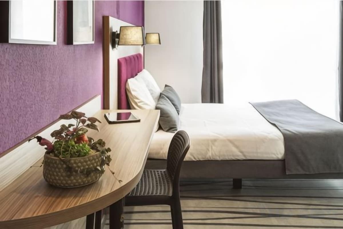 Une chambre d’hôtel avec un lit, une table de chevet avec une lampe, une chaise et une petite plante en pot.