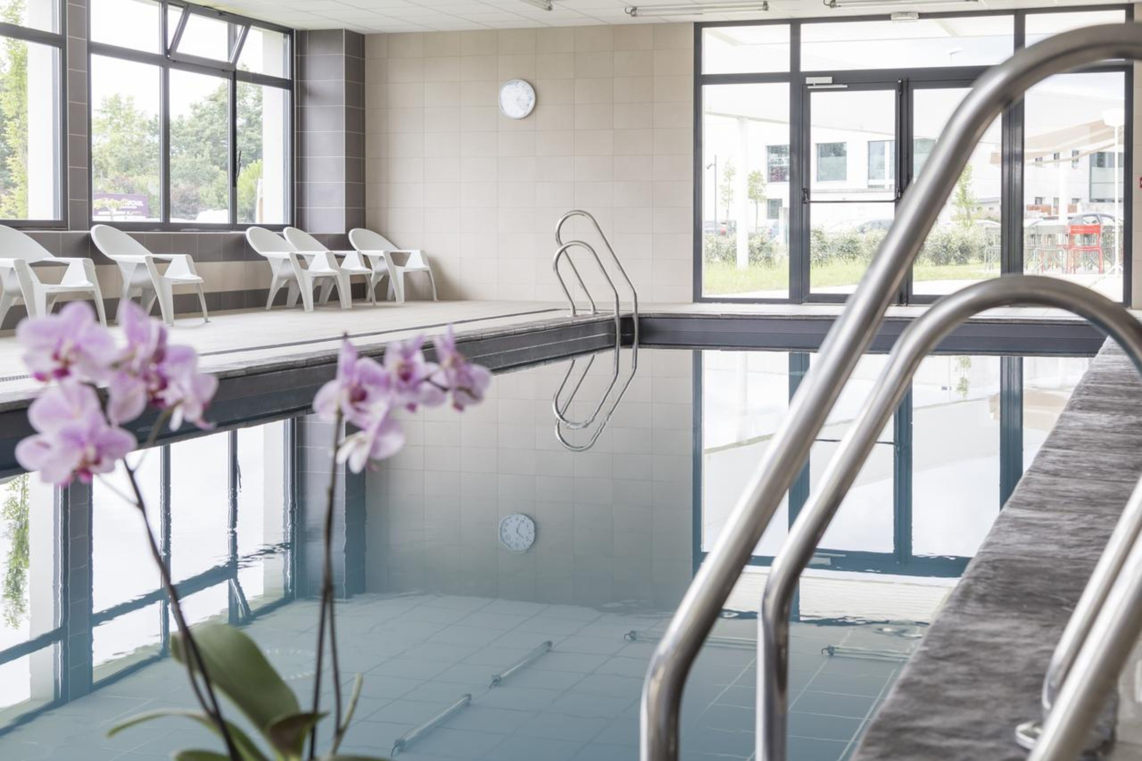 A modern indoor swimming pool with clear glass walls, a metal ladder, and white lounge chairs.