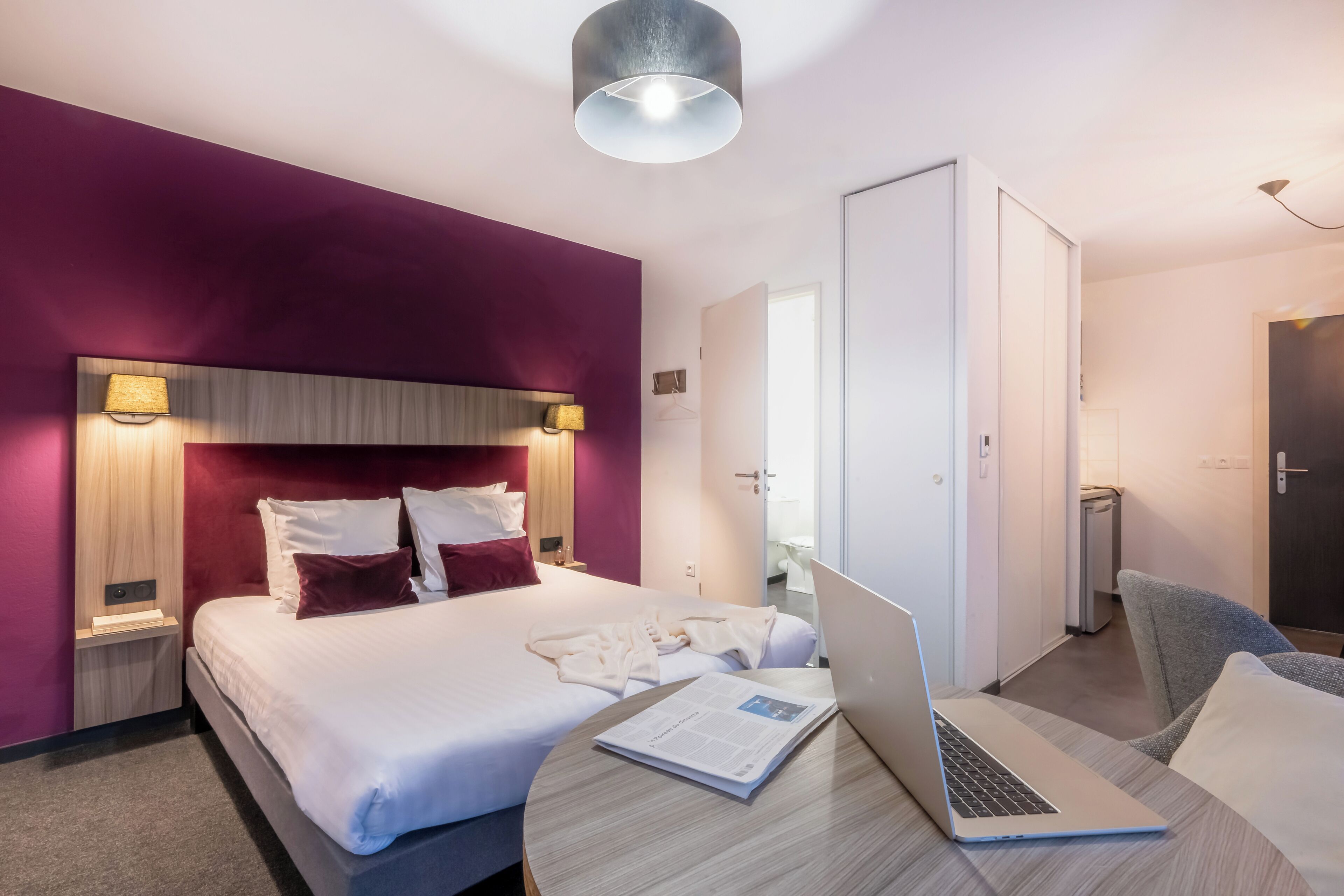 Een moderne hotelkamer met een groot bed, een laptop op een houten tafel en een deur naar een badkamer.