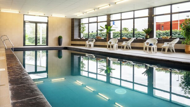 Une piscine intérieure avec des parois en verre transparent, des chaises blanches et des plantes en pot.