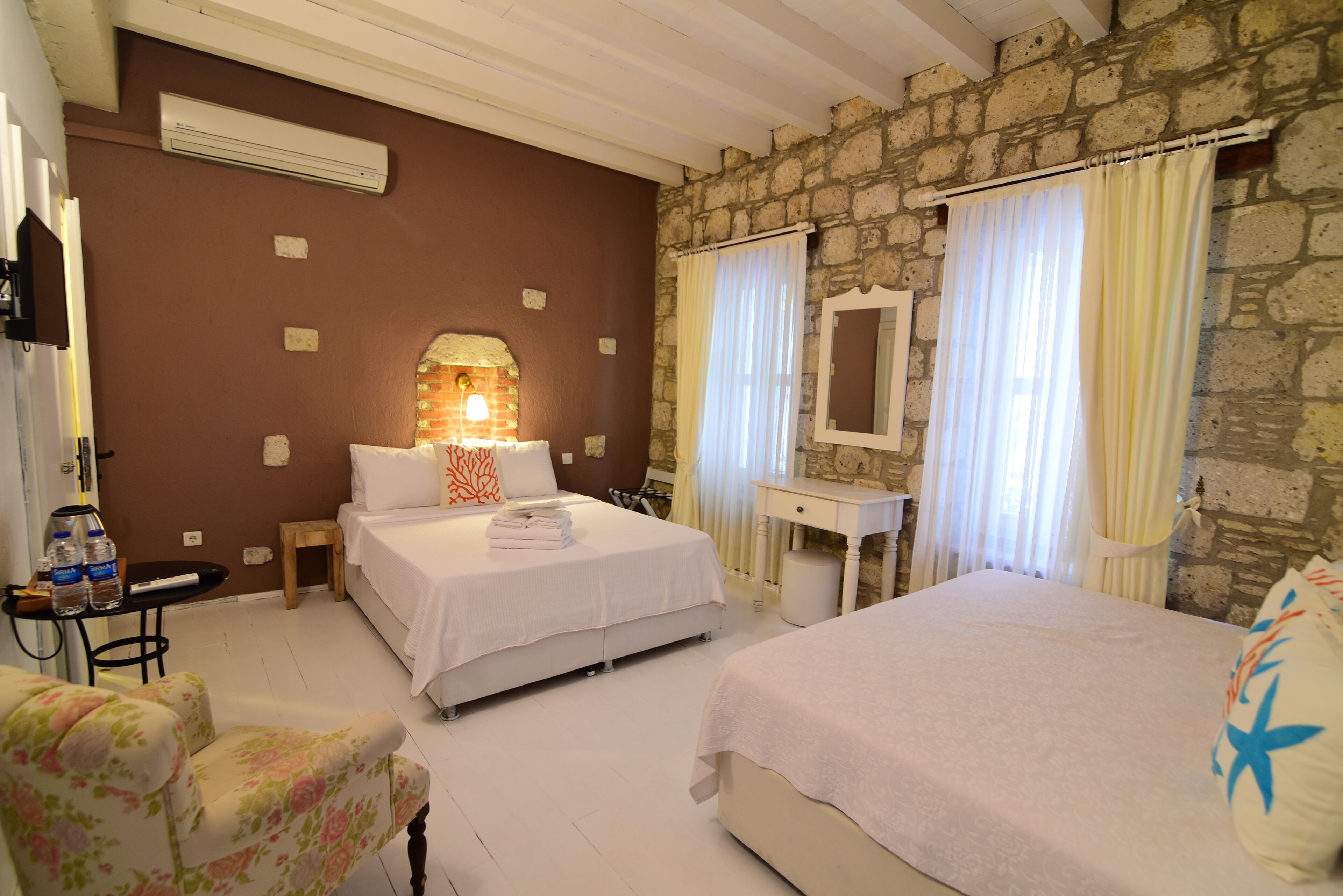 A hotel room with two beds, a desk, a chair, and a stone wall.