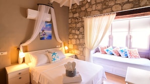 Un dormitorio con pared de piedra, cama con ropa de cama blanca, mesita de noche con lámpara y ventana con cortinas.