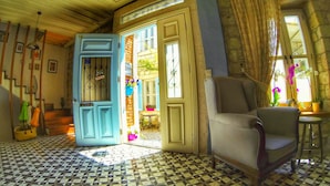 Una habitación con piso estampado, una puerta azul, un sillón gris y una ventana con cortinas.