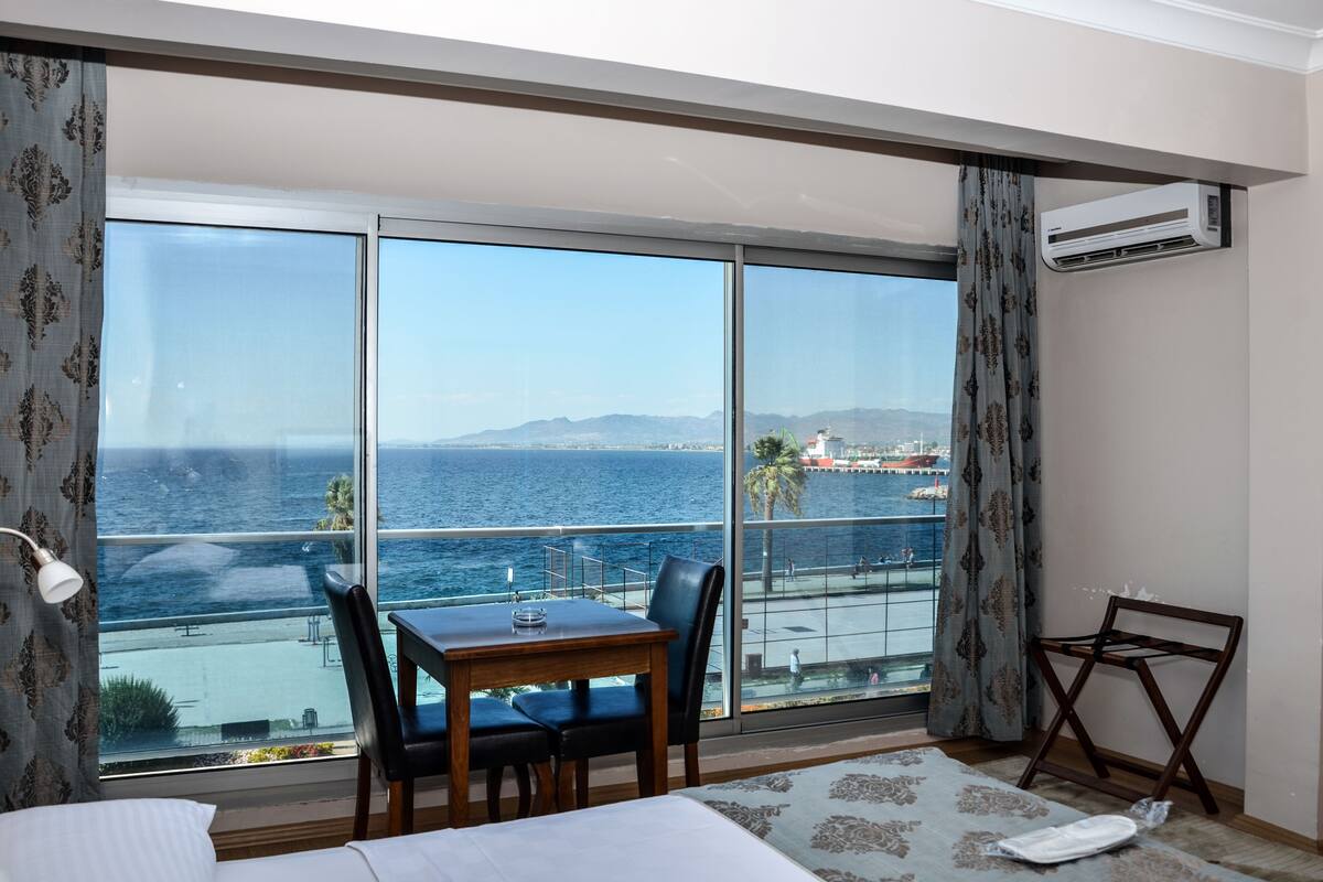 Habitación de hotel con un amplio ventanal corredero que da a un balcón con vistas al mar y montañas distantes.