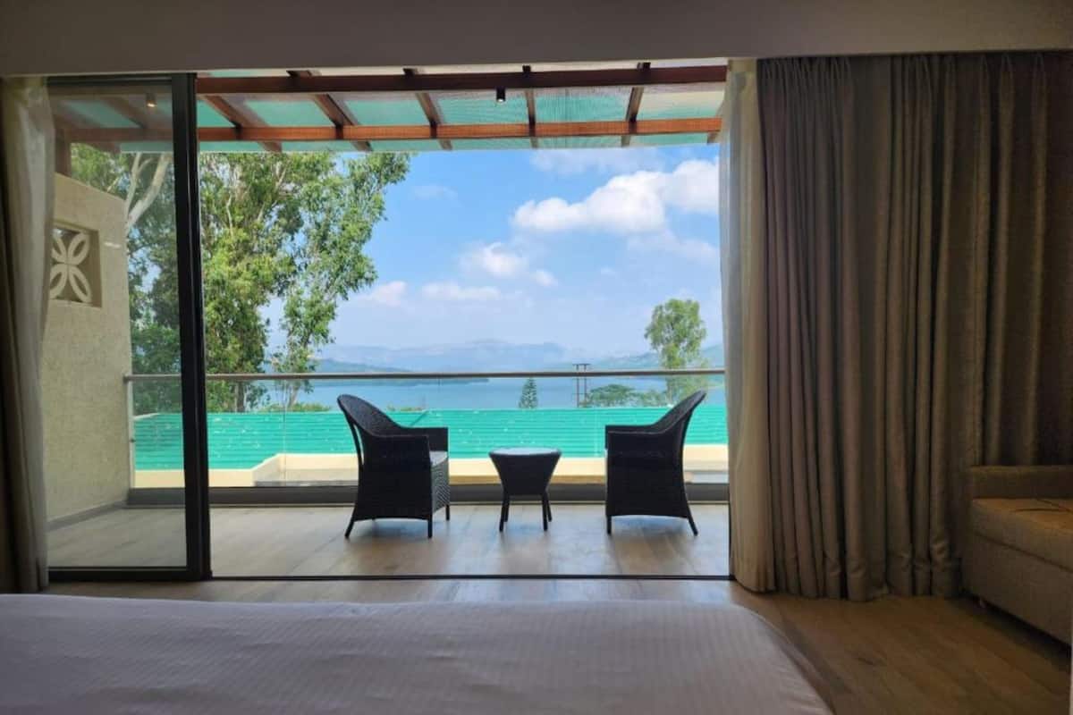 A bedroom with a large bed, a sliding glass door leading to a balcony with two chairs and a table, and a view of a body of water and greenery.