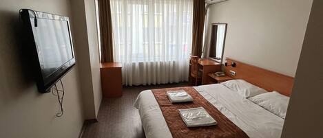 Una habitación de hotel con cama, escritorio, silla, televisor y ventana con cortinas.