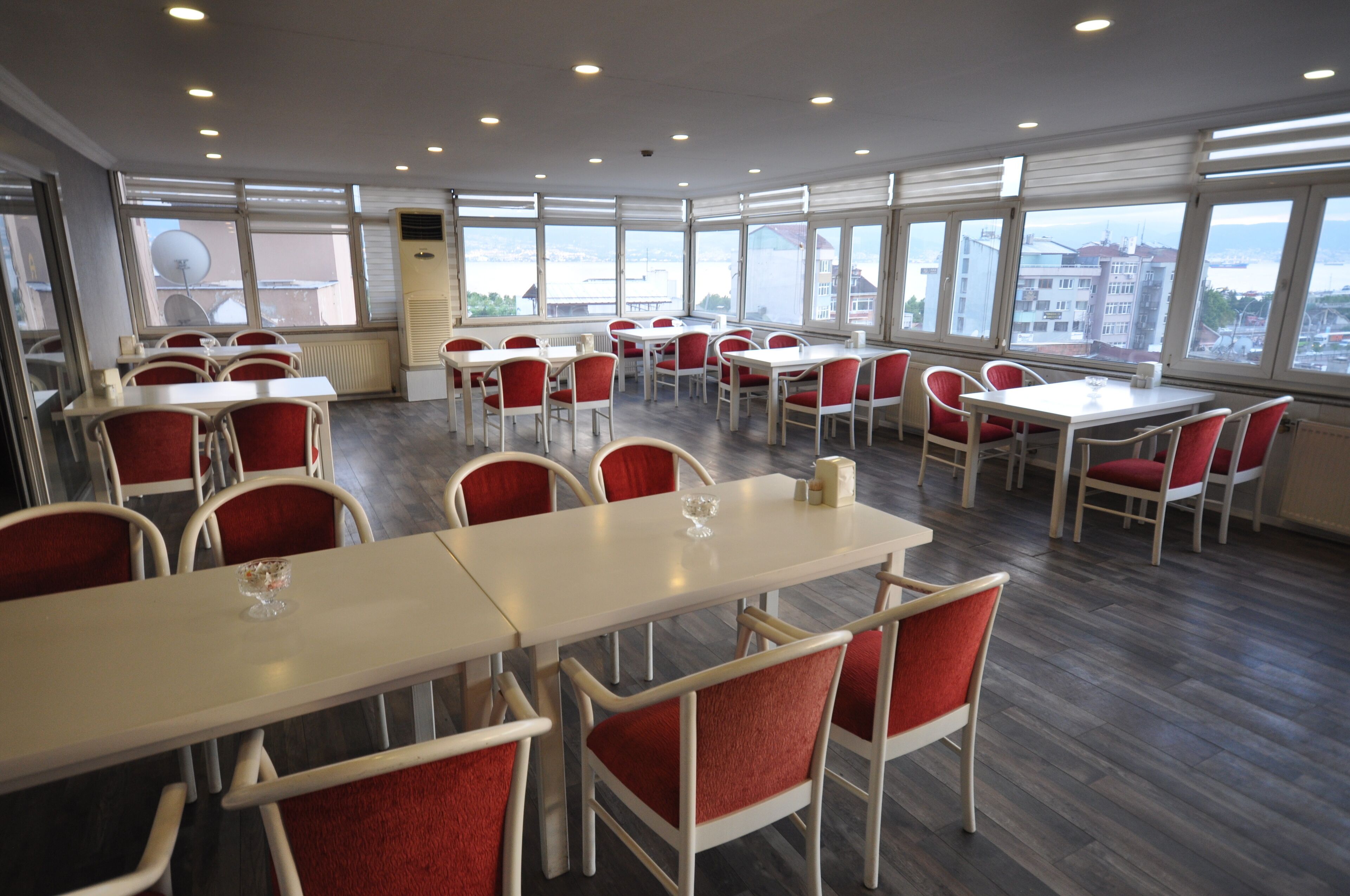 A spacious dining area with multiple tables and chairs, large windows offering a view of the cityscape, and a central air conditioning unit.