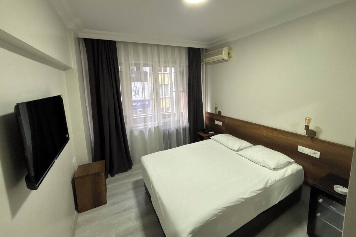 Une chambre d’hôtel avec un lit, un téléviseur à écran plat fixé au mur, une table de chevet avec une lampe et une fenêtre avec des rideaux.
