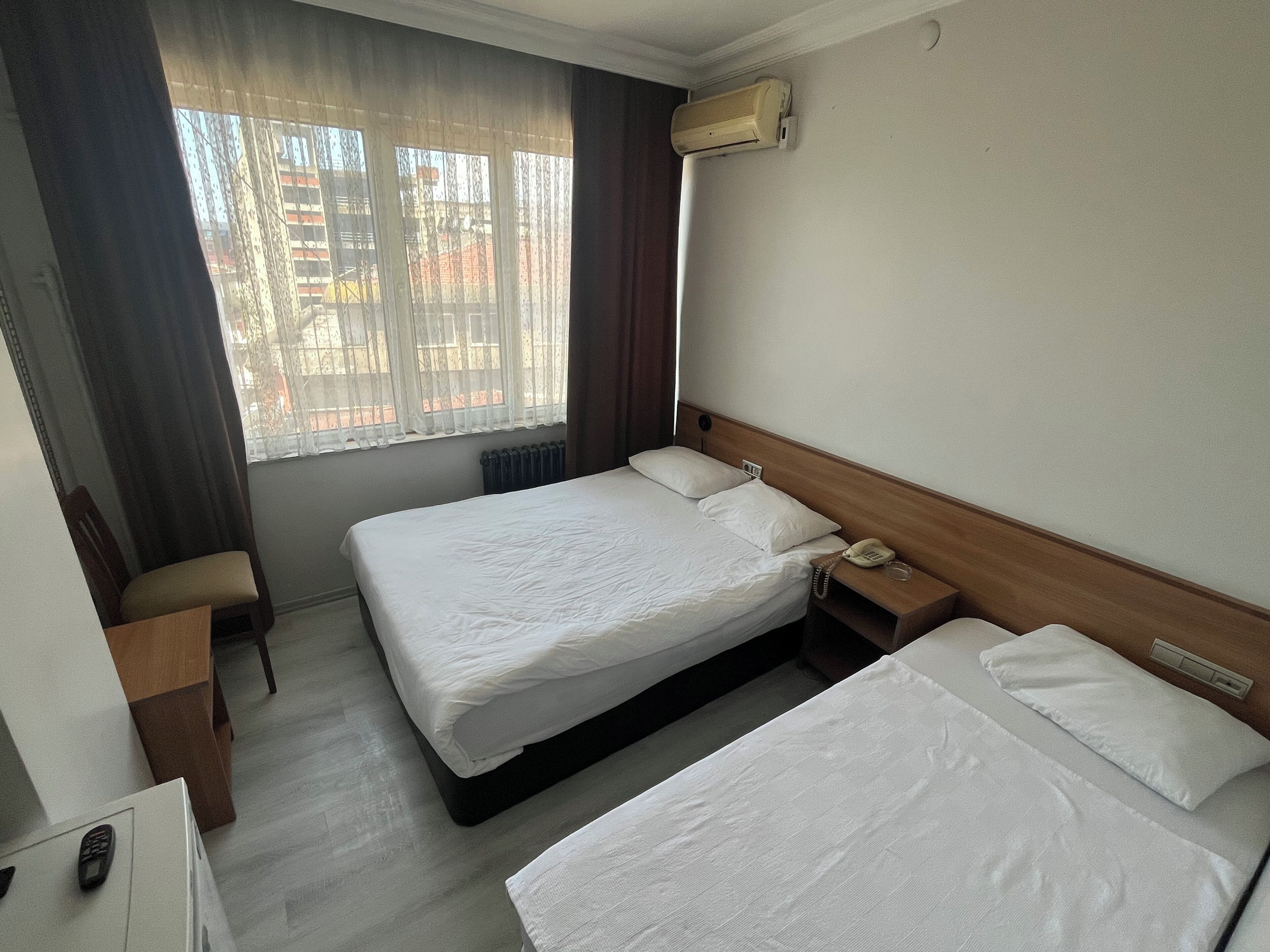 A hotel room with two beds, a bedside table with a phone, a radiator, and a window with curtains.