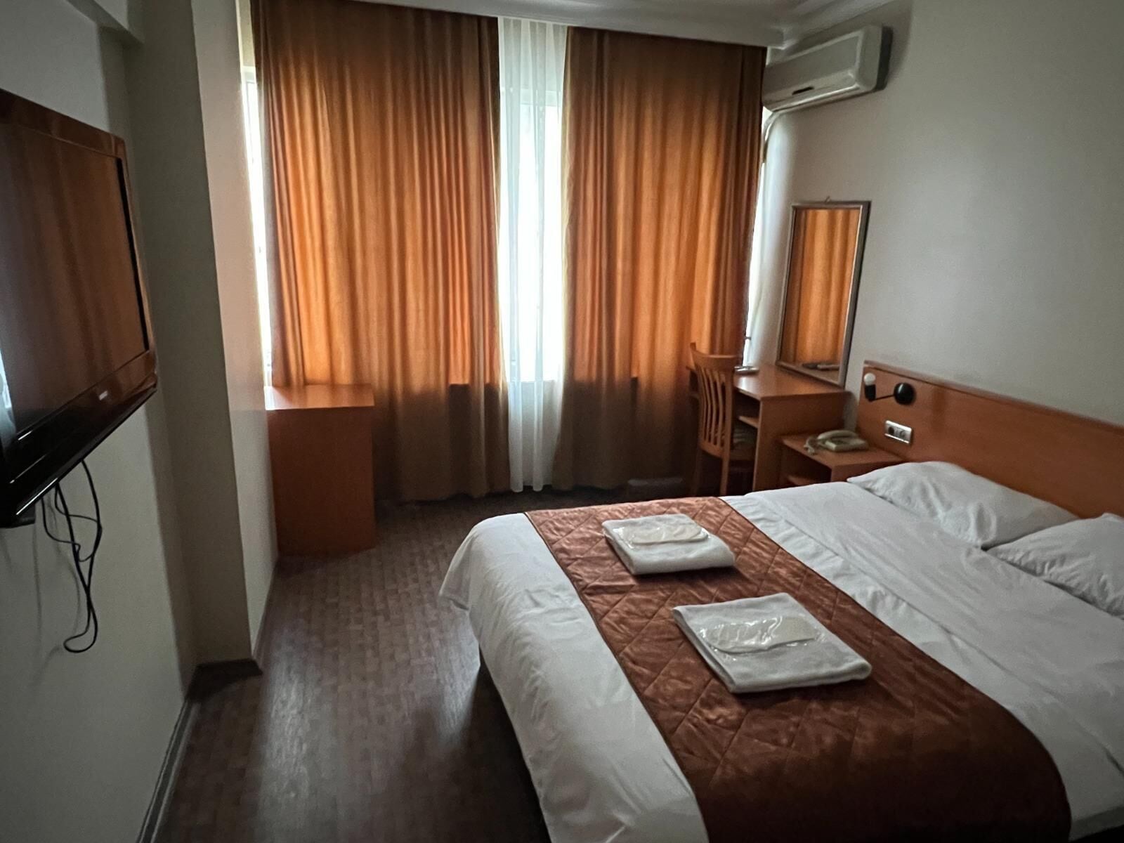 Quarto de hotel com cama, escrivaninha, cadeira, televisão e janela com cortinas.