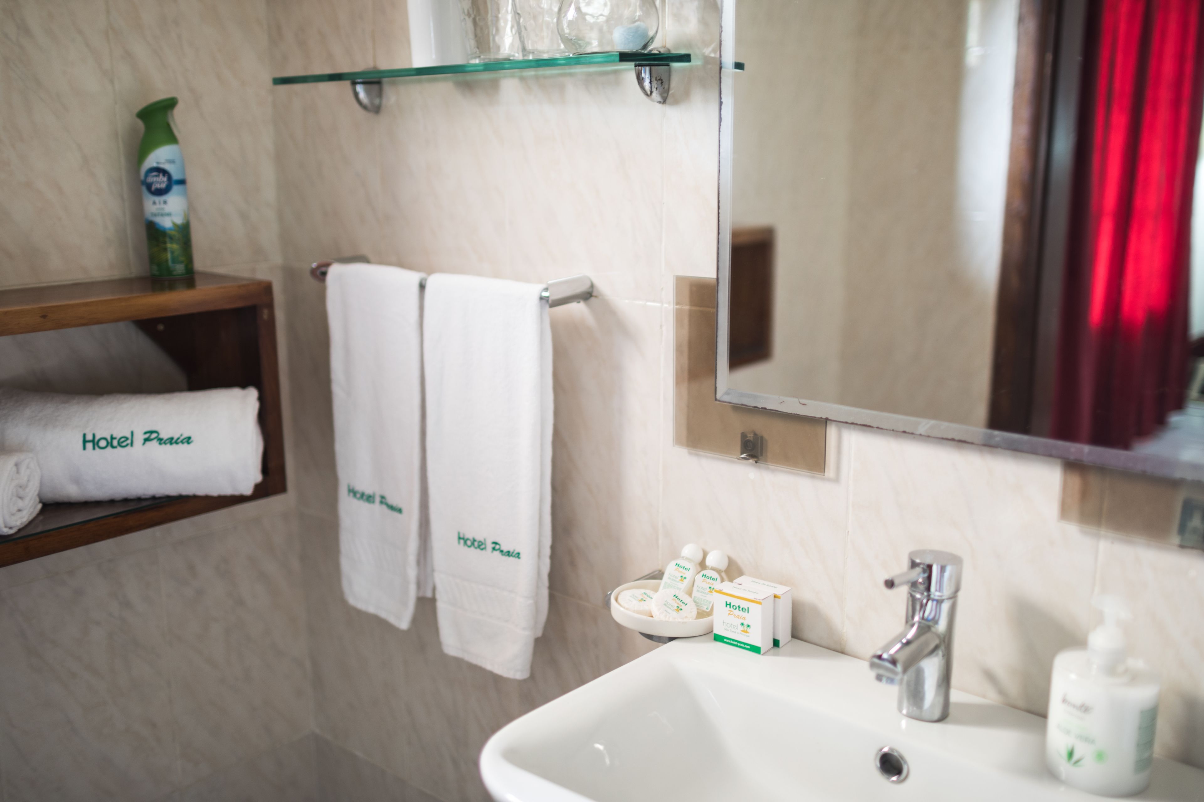 A bathroom with a white sink, a mirror, and a shelf with a bottle of shampoo and a towel labeled "Hotel Praia".
