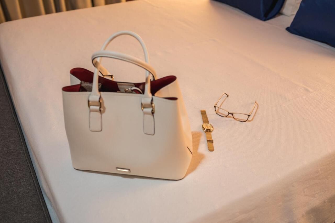 A white handbag, a pair of glasses, and a wristwatch on a bed.