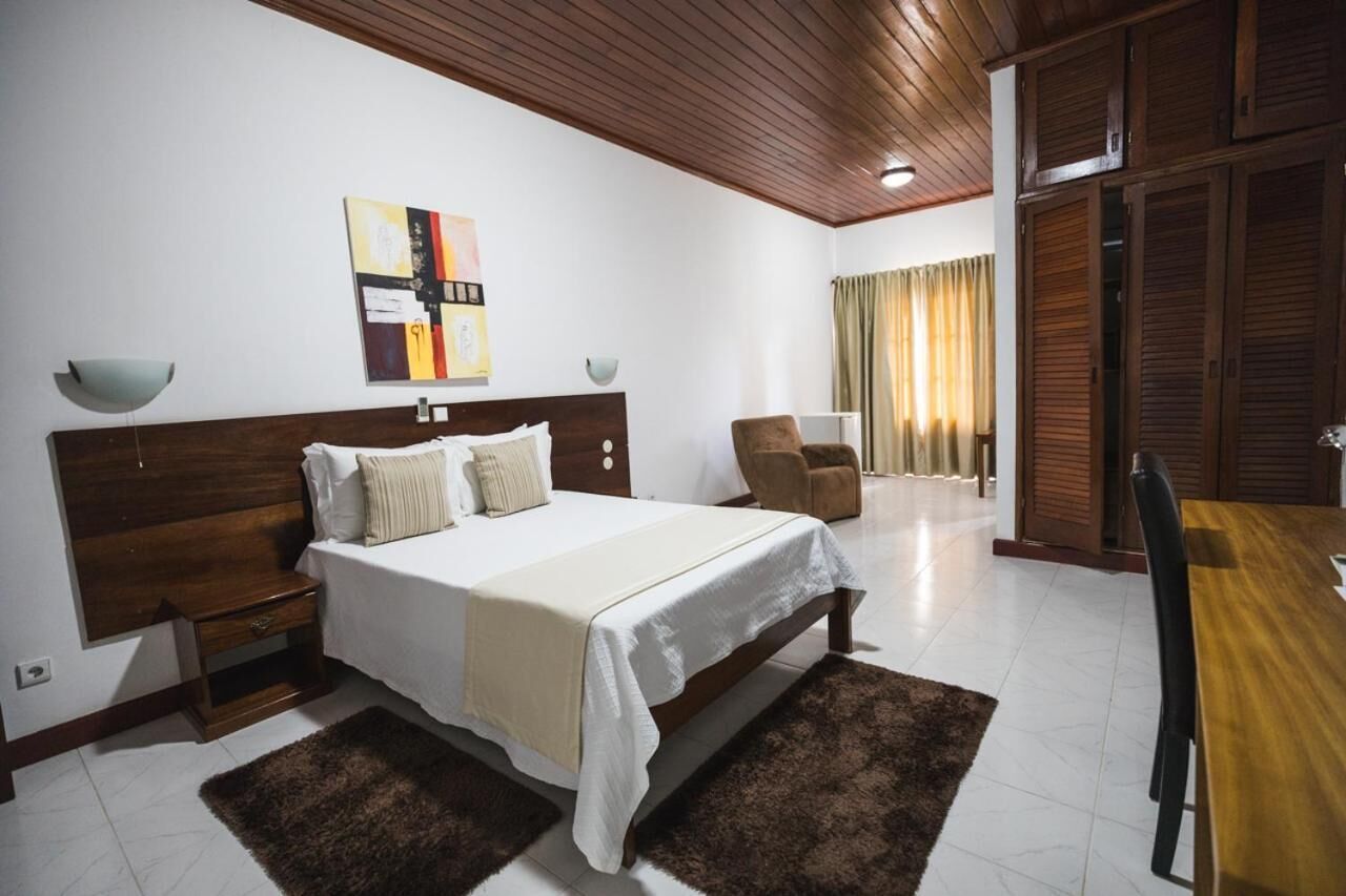 A bedroom with a wooden bed, a nightstand, a chair, a table, and a painting on the wall.