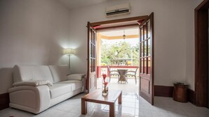 A living room with a sofa, a coffee table, and a red rug. There is a door leading to an outdoor area with a table and chairs. - Hotel Praia (São Tomé)