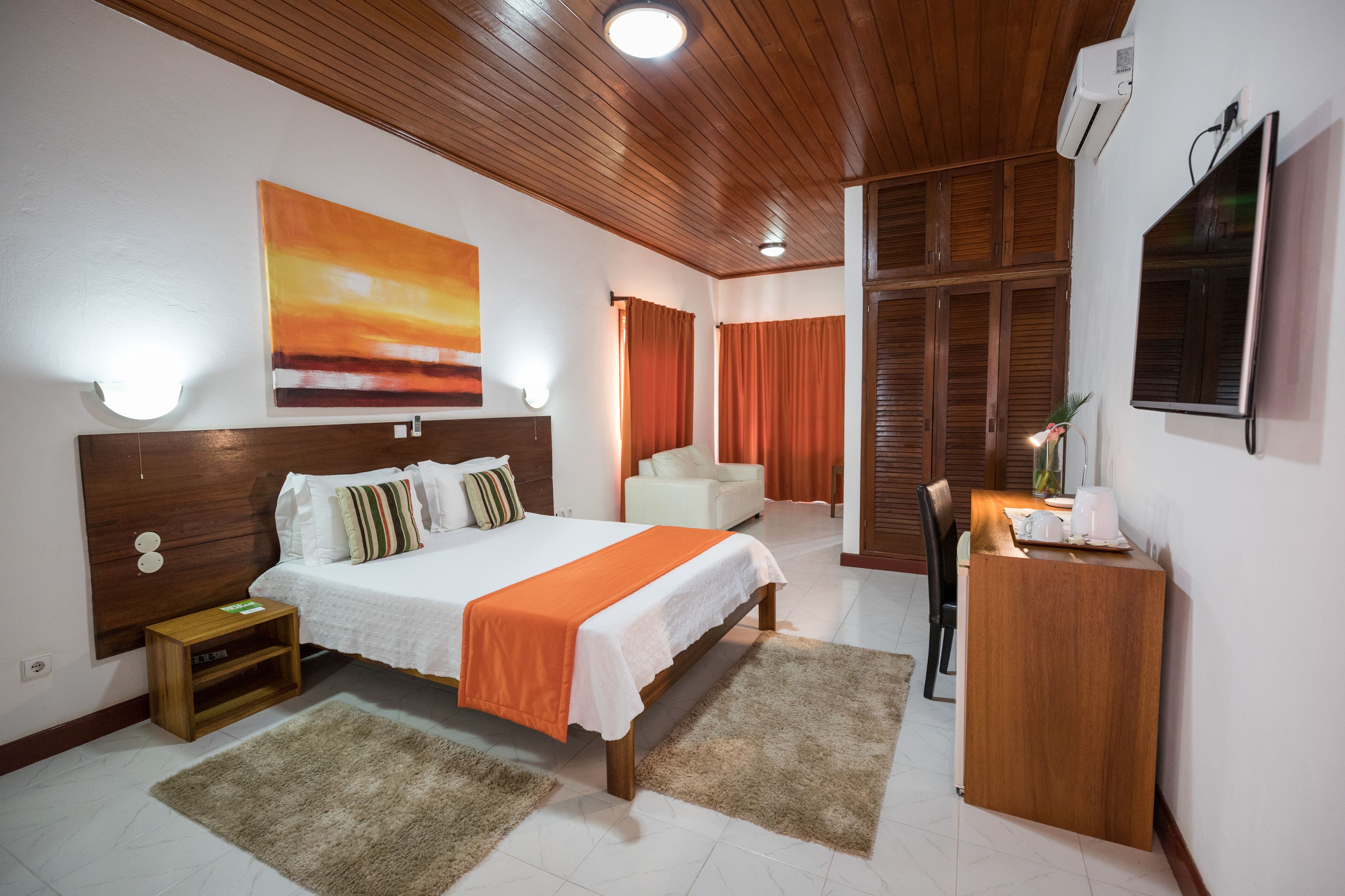 A hotel room with a bed, bedside tables, a desk, a chair, a TV, and a painting on the wall.