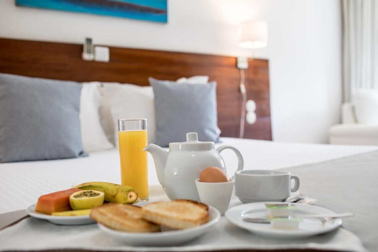 Una mesa de desayuno ordenada con tostadas, fruta, un calentador de agua, una taza y un vaso de zumo de naranja sobre una cama de hotel.