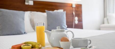 Una mesa de desayuno ordenada con tostadas, fruta, un calentador de agua, una taza y un vaso de zumo de naranja sobre una cama de hotel.
