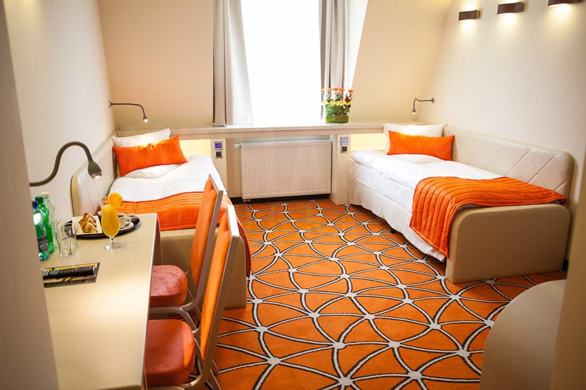 A hotel room with two beds, a desk, and a chair. There is a window with curtains, a radiator, and a small table with a glass of orange juice and a bottle of water.