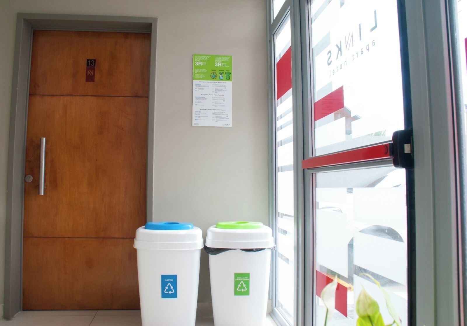 Uma porta de madeira com maçaneta, duas lixeiras brancas com símbolos de reciclagem e uma janela com toldo listrado em vermelho e branco.