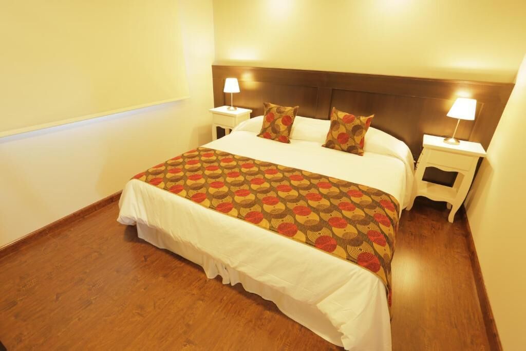 Une chambre d’hôtel avec un grand lit, des rideaux blancs, une couvre-lit à motifs, deux tables de chevet avec des lampes et un plancher en bois.