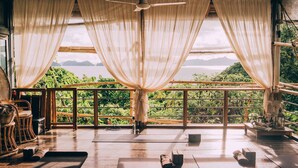 A room with a wooden floor, a ceiling fan, and a view of a lake and mountains through sheer curtains. - The Birdhouse El Nido - Glamping (El Nido)