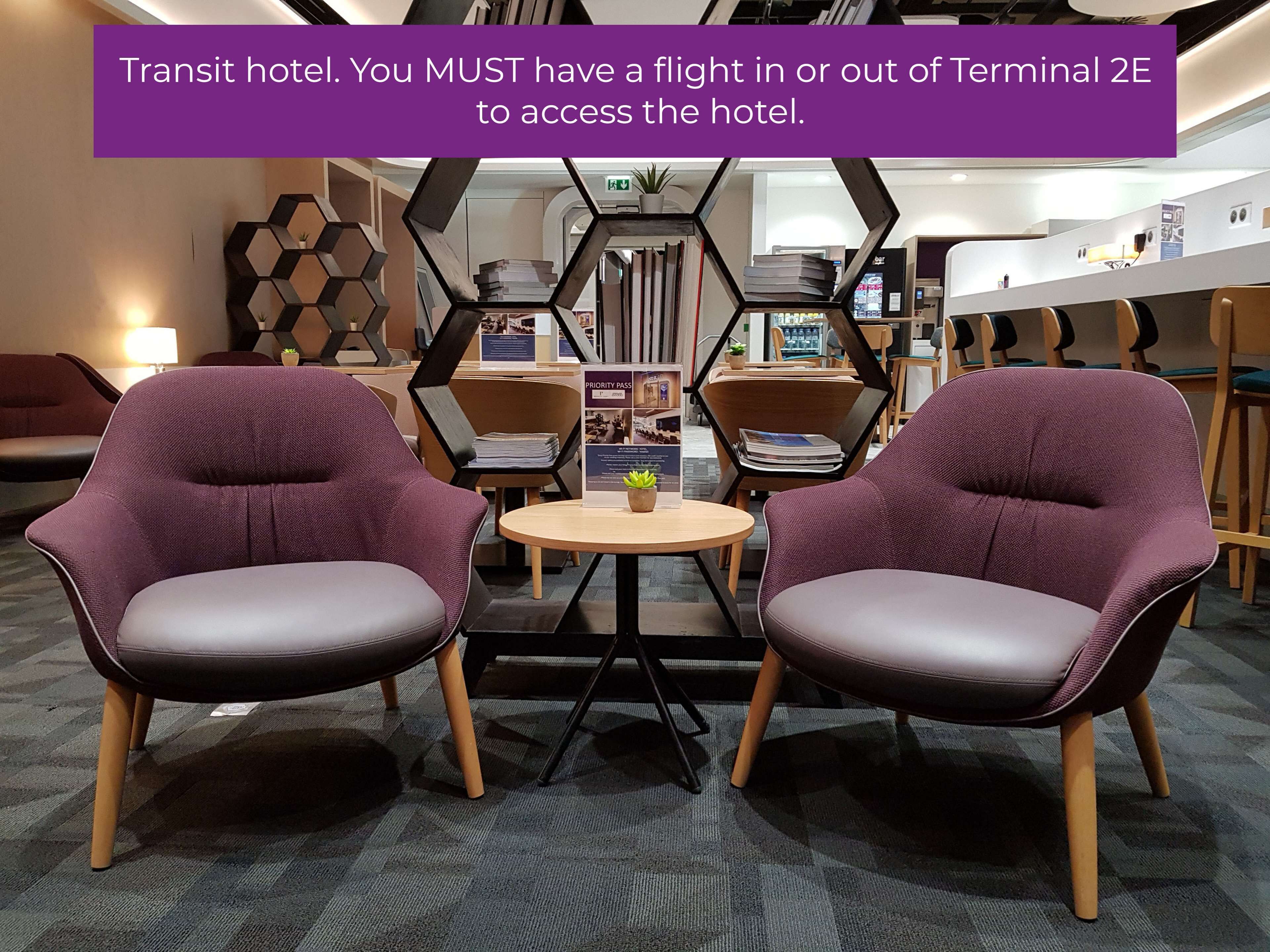 A modern hotel lobby with two purple armchairs, a small round table, and a hexagonal bookshelf.
