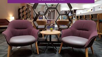 A modern hotel lobby with two purple armchairs, a small round table, and a hexagonal bookshelf.