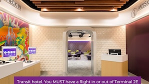 Ein Empfangsbereich eines Transit-Hotels mit einer Rezeption, einem farbenfrohen Wandgemälde und einer sichtbaren Sitzecke durch eine Glastür.