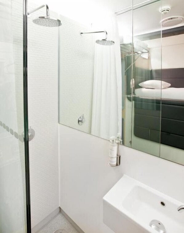A modern bathroom with a glass shower enclosure, a white sink, and a large mirror reflecting a bed.
